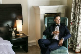 Ein Bräutigam, der entspannt in einem eleganten Sessel in der Hochzeitssuite des Hotel Atlantic in Hamburg sitzt. Er trägt einen schicken Anzug und hält ein Glas Whiskey in der Hand. Der exklusive Stil der Suite, kombiniert mit dem entspannten Auftreten des Bräutigams, vermittelt ein Gefühl von Eleganz und Gelassenheit.