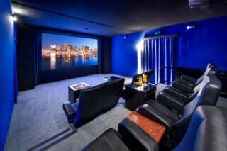 Ein privates Kino, das für besondere Anlässe gemietet werden kann. In dem stilvoll eingerichteten Raum befinden sich gemütliche Kinosessel, die zu einem entspannten Filmerlebnis einladen. Auf den Tischen vor den Sitzen stehen elegant garnierte Cocktails, die den exklusiven Charakter des Kinos unterstreichen. Die warme Beleuchtung und die luxuriöse Einrichtung schaffen eine einladende Atmosphäre für ein perfektes Kinoerlebnis in privater Umgebung.






