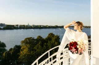 Hotel Atlantic Eine Braut, die in einem eleganten weißen Kleid auf einem Balkon des Hotel Atlantic in Hamburg steht. Sie hält einen wunderschönen Brautstrauß in den Händen. Im Hintergrund ist die sonnenbeschienene Alster zu sehen, die eine malerische Kulisse für diesen besonderen Moment bietet. Die Braut blickt in die Ferne, während das Sonnenlicht ihr Kleid und den Balkon in ein warmes, goldenes Licht taucht. Die Szene strahlt Romantik und Eleganz aus und fängt die Schönheit des Augenblicks perfekt ein.