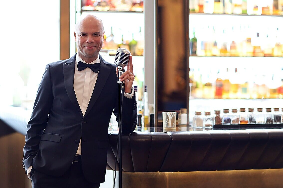 Mann im Smoking mit Mikrofon vor einer Bar mit verschiedenen Getränken im Hintergrund.