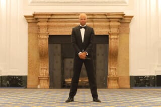 Dennis Durant steht im Black Tie Outfit vor dem Kamin des Großen Festsaals und schaut ernsthaft in die Kamera.