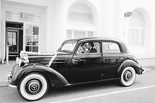 Das Schwarzweiß-Bild zeigt ein Hochzeitspaar in einem eleganten schwarzen Mercedes aus den 1940er Jahren. Das Paar sitzt im Auto und küsst sich zärtlich. Der klassische Wagen und die Schwarzweiß-Fotografie verleihen der Szene einen nostalgischen und zeitlosen Charme.