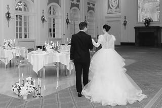 Ein frisch verheiratetes Paar, das von hinten zu sehen ist. Der Bräutigam trägt einen eleganten schwarzen Anzug, während die Braut ein wunderschönes weißes Kleid mit langer Schleppe trägt. Das Paar schreitet Hand in Hand durch einen festlich geschmückten Hochzeitssaal. Die Tische im Saal sind mit eleganten Dekorationen und Blumenschmuck versehen, was die festliche und romantische Atmosphäre der Hochzeitsfeier unterstreicht.