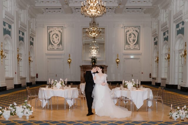 Ein frisch verheiratetes Ehepaar, das bei ihrer Hochzeit in einem prachtvoll geschmückten historischen Saal des Hotel Atlantic Hamburg steht. Das Paar befindet sich in der Mitte des Raumes und wird von der eleganten Atmosphäre des Saals umgeben. Auf dem Boden sind wunderschöne Blumensträuße arrangiert, die zur festlichen Dekoration beitragen. Die Tische im Hintergrund sind exquisit gedeckt und bereit für die Hochzeitsgesellschaft.