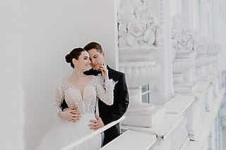 Ein Hochzeitspaar, das in einem eleganten Gang mit weißen Säulen steht. Der Bräutigam steht hinter der Braut und hält liebevoll ihre Hand. Die Braut hat sich zu ihm umgedreht und lächelt ihn glücklich an. Die weißen Säulen und die elegante Architektur des Gangs verleihen der Szene eine klassische und zeitlose Atmosphäre, die perfekt die Romantik und den Zauber dieses besonderen Moments einfängt.