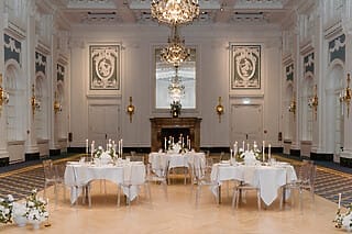 Eine historischer, eleganter und exklusiver Hochzeitssalon, der durch einen beeindruckenden Kronleuchter und die prachtvolle Dekoration besticht. Die Tische sind festlich gedeckt und bereit für die Gäste, mit feinem Geschirr und eleganten Kerzenhaltern. Auf jedem Tisch brennen Kerzen, die eine warme und romantische Atmosphäre schaffen. Der Saal strahlt Luxus und Raffinesse aus, und unterstreicht die Exklusivität des Ortes, ideal für eine unvergessliche Hochzeitsfeier.