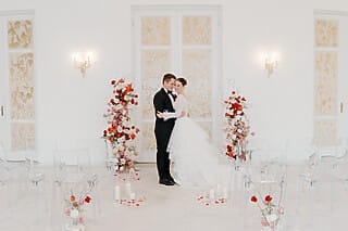 Das Bild zeigt ein Hochzeitspaar in einem hellen Saal, der festlich für die Zeremonie dekoriert ist. Links und rechts des Paares sind wunderschöne Rosenarrangements platziert, die den romantischen Charakter des Anlasses betonen. Auf beiden Seiten des Gangs stehen elegant arrangierte Stühle für die Gäste, die darauf warten, die Zeremonie mitzuerleben. Der helle Saal, gefüllt mit natürlichem Licht und floralen Akzenten, schafft eine einladende und festliche Atmosphäre für diesen besonderen Moment.