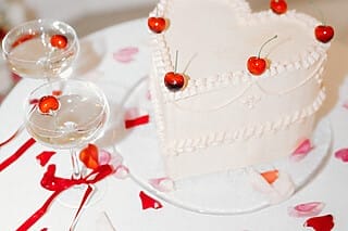 Eine wunderschöne Hochzeitstorte, die mit frischen Kirschen dekoriert ist. Daneben stehen zwei Cocktails, die ebenfalls mit Kirschen garniert sind, was ein harmonisches Bild ergibt. Die Kombination von Torte und Cocktail unterstreicht die Liebe zum Detail und die sorgfältige Planung der Hochzeitsfeier.
