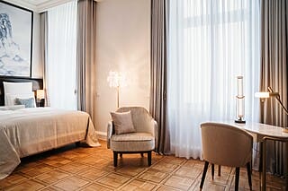 Elegantes Hotelzimmer mit Bett, Sessel und Schreibtisch, hellen Vorhängen und stilvoller Beleuchtung.