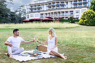 Falkenstein Grand Ein Mann und eine Frau genießen ein Picknick in einem sonnigen Park. Sie sitzen auf einer Decke, umgeben von einem Picknickkorb und einer Flasche Sekt.