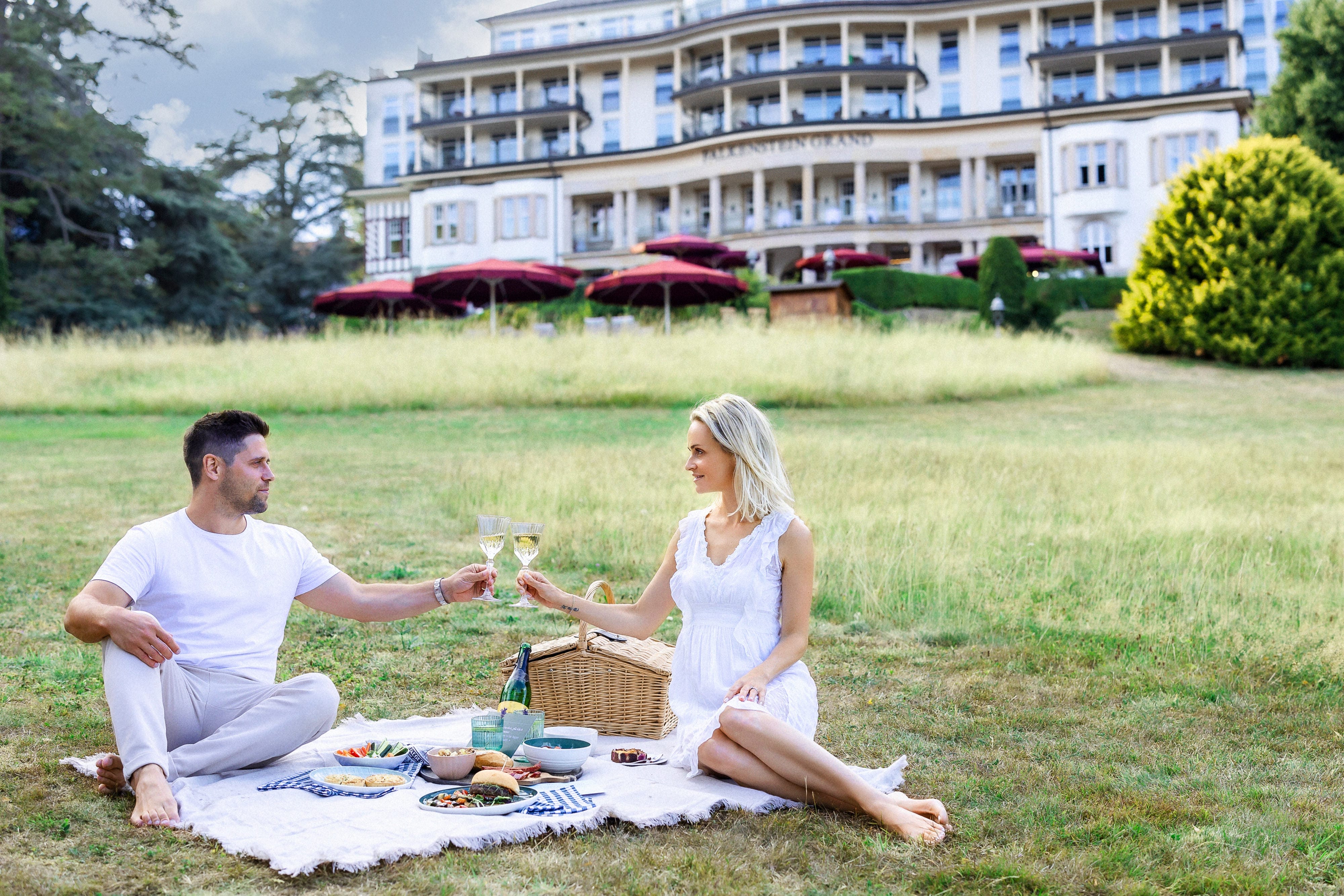 Falkenstein Grand Ein Mann und eine Frau genießen ein Picknick in einem sonnigen Park. Sie sitzen auf einer Decke, umgeben von einem Picknickkorb und einer Flasche Sekt.