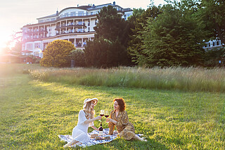 Falkenstein Grand Ein Picknick bei Sonnenuntergang auf einer Wiese im Park, zwei Personen stoßen freudig mit Rotweingläsern an, genießen die sommerliche Atmosphäre.