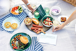 Falkenstein Grand Eine Picknickdecke mit mediterraner Quiche, Antipasti, Gemüsesnacks und saftigen Beef-Burgern, bereit für ein gemütliches Picknick im Freien.