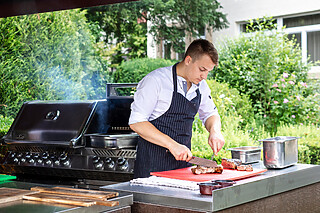 Falkenstein Grand Skyline BBQ in Frankfurt. Ein Koch steht im Park neben einem Barbeque-Grill und bereitet Fleisch vor, während der Rauch vom Grill in die Luft aufsteigt.